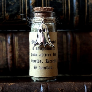 May include: A small glass bottle with a cork stopper and a silver ghost charm. The bottle is filled with a brown powder and has a label that reads "Po utel a saupoudrer pour attirer les esprits. Recellt de bandou."