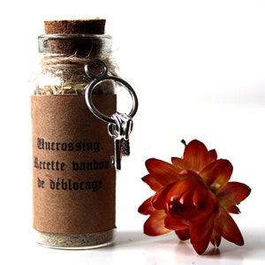 Può includere: Una piccola bottiglia di vetro con tappo di sughero, riempita di erbe essiccate e un portachiavi d'argento. La bottiglia ha un'etichetta marrone con il testo "Uncrossing. Recette vaudou de déblocage." Un fiore arancione secco è accanto.