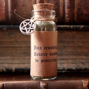 Può includere: Una piccola bottiglia di vetro con un tappo di sughero e un'etichetta di carta marrone. L'etichetta riporta il testo "Jinx removing Recette vaudois de protection". Un ciondolo a pentagramma d'argento è appeso alla bottiglia.