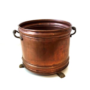 May include: A cylindrical copper vessel with a polished metallic finish, featuring two small curved side handles and three decorative feet at the base. The container has a wide open top and a smooth, rounded rim, showcasing a warm reddish-brown tone.