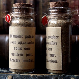 Può includere: Due piccole bottiglie di vetro piene di polvere marrone, ciascuna sigillata con un tappo di sughero e spago. Le bottiglie hanno etichette con testo francese e sono adornate da un ciondolo rosso. Su uno sfondo di libri antichi.