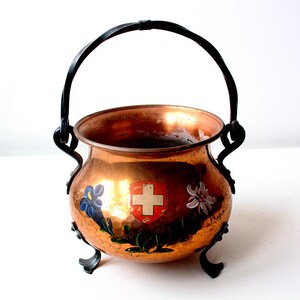 May include: A copper pot with a black handle and three legs. The pot is decorated with a Swiss flag and flowers.