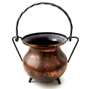 May include: A hammered copper cauldron with a black twisted handle and three small legs. The cauldron has a dark interior and is designed with a rounded body and a wide rim. The handle is attached to the cauldron with two metal supports.