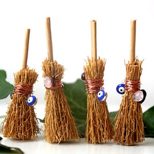 Könnte beinhalten: Vier Miniaturbesen mit Holzstielen und natürlichen Faserborsten. Jeder Besen ist mit Kupferdraht und Charms verziert, darunter blaue Glücksbringer-Perlen und rosa und braune Perlen.
