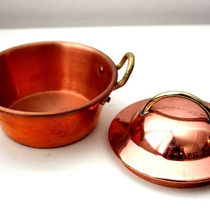 May include: A small copper pot with two brass handles and a matching copper lid with a brass handle. The pot and lid are shiny and reflect light.