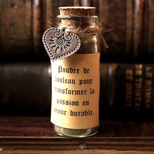 Può includere: Una piccola bottiglia di vetro con un tappo di sughero e un'etichetta di carta marrone. L'etichetta ha un testo in francese che recita "Poudre de bouleau pour transformer la passion en amour durable." La bottiglia è legata con uno spago e ha un ciondolo a forma di cuore argentato.