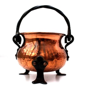 Pode incluir: Uma pequena panela redonda de cobre martelado com uma alça de metal escuro torcido e três pés de metal escuro correspondentes. A panela tem uma abertura larga e uma superfície metálica reflexiva com marcas visíveis do martelamento.