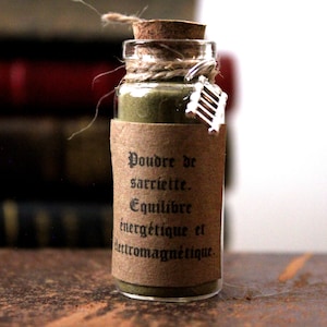 Peut inclure: Petite bouteille en verre remplie de poudre verte, scellée avec un bouchon de liège et de la ficelle. Une étiquette en papier brun avec du texte noir indique "Poudre de sarriette. Equilibre énergétique et électromagnétique." Un petit charme argenté est suspendu à la ficelle.