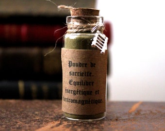 Pode incluir: Uma pequena garrafa de vidro cheia de pó verde, selada com uma rolha e barbante. Um rótulo de papel castanho com texto preto diz "Poudre de sarriette. Equilibre énergétique et électromagnétique." Um pequeno pingente prateado pende do barbante.