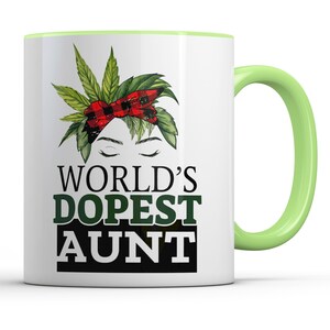 May include: White ceramic mug with a green and red marijuana leaf design and the text "WORLD'S DOPEST AUNT". The mug has a light green rim and handle.