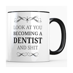 May include: White ceramic coffee mug with a black rim and handle. The mug has a black design that says "Look at you becoming a dentist and shit" with decorative swirls above and below the text.