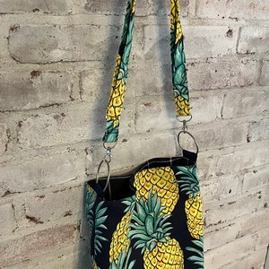 Peut inclure: Un sac cabas noir avec un imprimé ananas. Le sac a une longue bandoulière et est fait d'un tissu robuste.