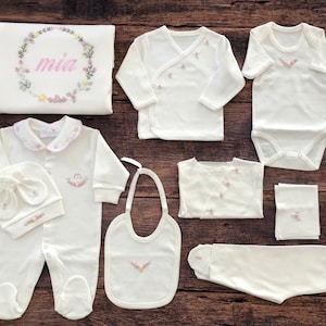 May include: A white baby outfit set with floral embroidery. The set includes a onesie, a hat, a bib, a pair of pants, a swaddle blanket, and two shirts. The blanket has the name "mia" embroidered on it.