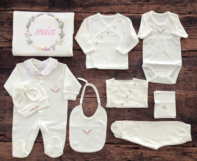 May include: A white baby outfit set with floral embroidery. The set includes a onesie, a hat, a bib, a pair of pants, a swaddle blanket, and two shirts. The blanket has the name "mia" embroidered on it.