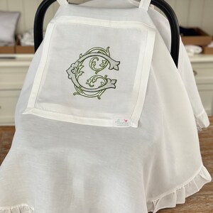 May include: White baby car seat cover with a ruffled edge and a monogrammed initial "C" in a green floral design. The cover has a small white tag with the word "Florrie" on it.