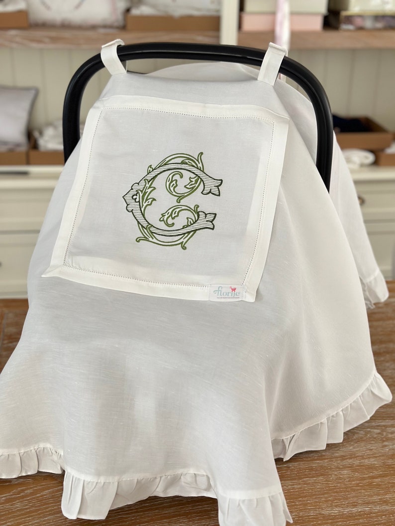 May include: White baby car seat cover with a ruffled edge and a monogrammed initial "C" in a green floral design. The cover has a small white tag with the word "Florrie" on it.