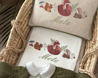 May include: A white baby onesie with a green cardigan and a white embroidered pillow with a fox, squirrel, hedgehog, and bird design. The pillow has the name "Mette" embroidered on it.