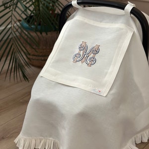 May include: White linen car seat cover with a ruffled edge and an embroidered letter "K" in a decorative font. The cover has a small "florie" label.