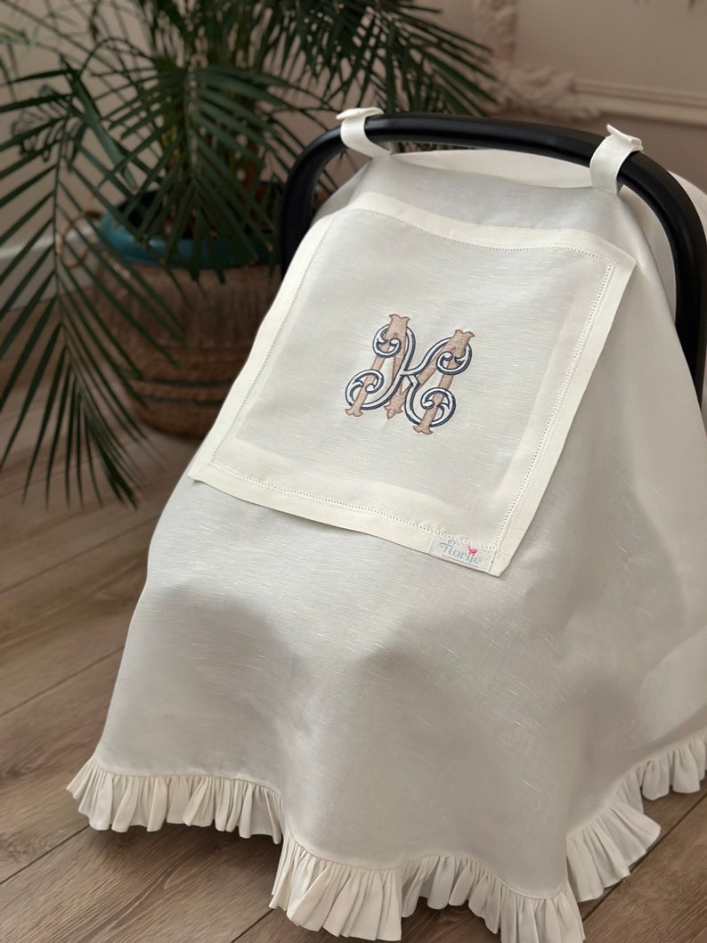 May include: White linen car seat cover with a ruffled edge and an embroidered letter "K" in a decorative font. The cover has a small "florie" label.