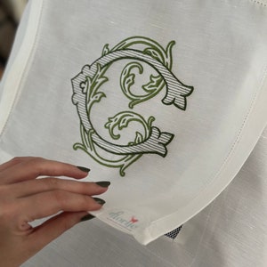 May include: White linen napkin with a green embroidered letter C in a decorative floral design. The napkin has a small pocket with the word "florie" printed on it.