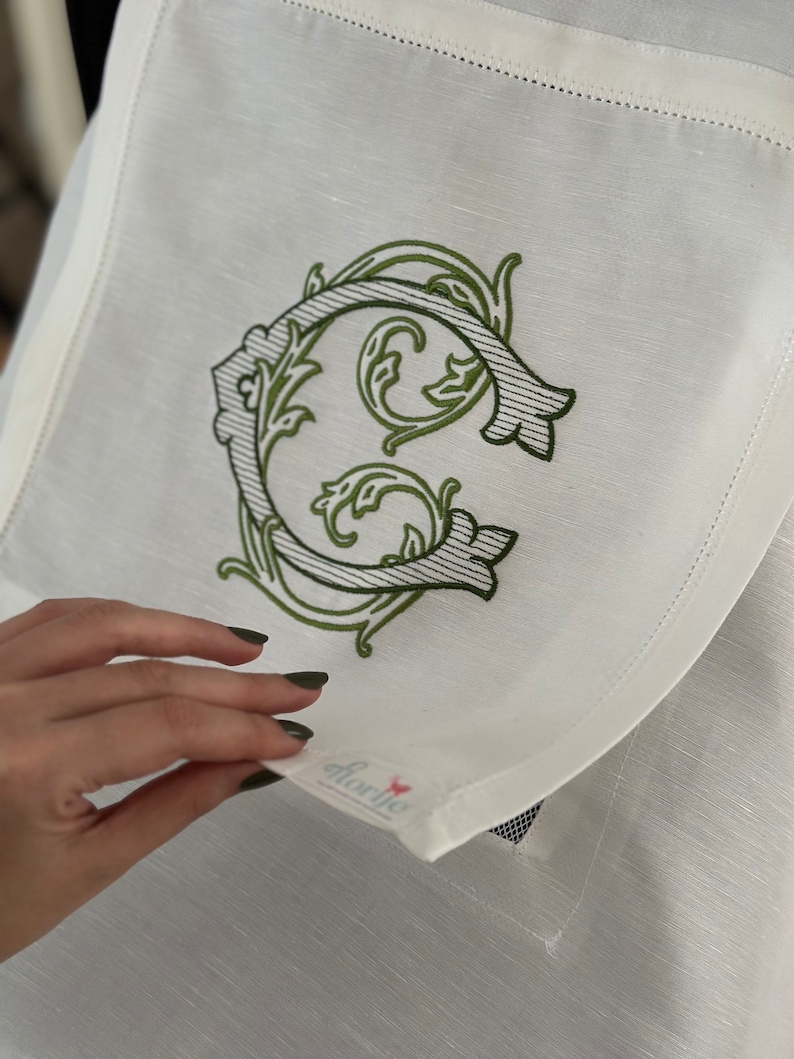 May include: White linen napkin with a green embroidered letter C in a decorative floral design. The napkin has a small pocket with the word "florie" printed on it.