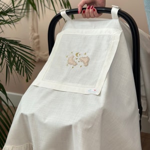 May include: White stroller cover with a ruffle trim and embroidered clouds, stars, and a crescent moon in gold thread. The cover has a small white tag with the word "Flamingo" in pink.