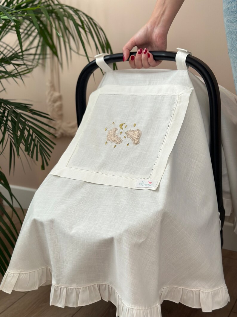 May include: White stroller cover with a ruffle trim and embroidered clouds, stars, and a crescent moon in gold thread. The cover has a small white tag with the word "Flamingo" in pink.