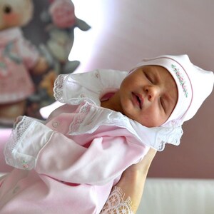 May include: A sleeping baby wearing a pink and white outfit with a white lace collar and a white hat with pink trim and floral embroidery.