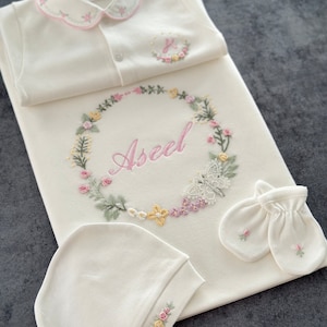 May include: A white baby outfit set with embroidered flowers and the name "Aseel" in pink lettering. The set includes a onesie, a blanket, a hat, and a pair of mittens.