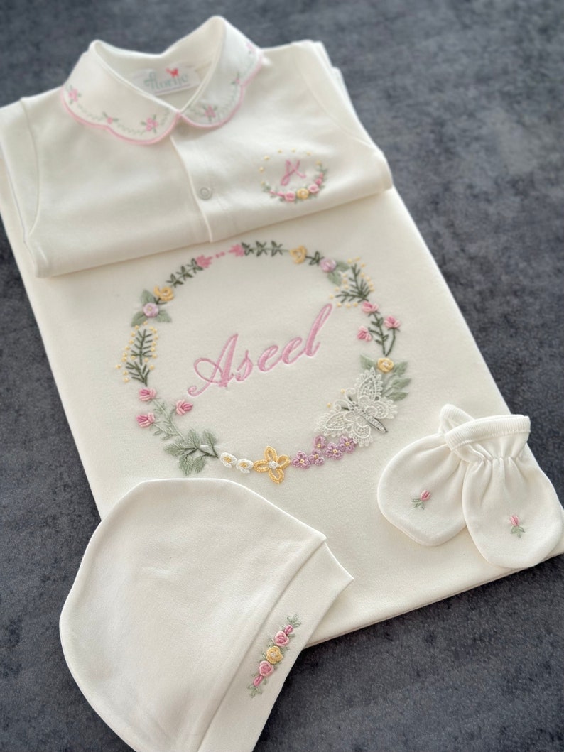 May include: A white baby outfit set with embroidered flowers and the name "Aseel" in pink lettering. The set includes a onesie, a blanket, a hat, and a pair of mittens.