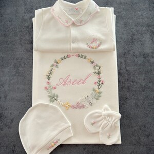 May include: A white baby outfit set with floral embroidery. The set includes a onesie, a hat, a pair of mittens, and a swaddle blanket. The onesie has a Peter Pan collar and a button closure. The swaddle blanket has a floral wreath with the name "Aseel" embroidered in pink. The set is perfect for a baby shower gift.