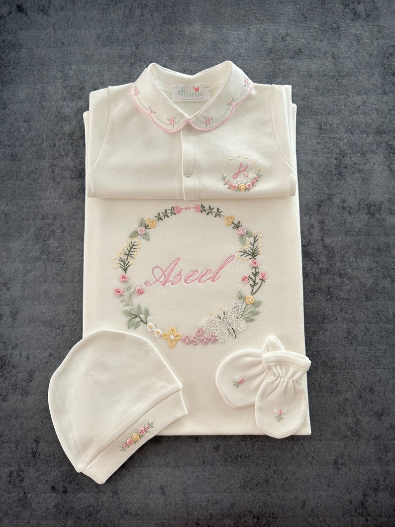 May include: A white baby outfit set with floral embroidery. The set includes a onesie, a hat, a pair of mittens, and a swaddle blanket. The onesie has a Peter Pan collar and a button closure. The swaddle blanket has a floral wreath with the name "Aseel" embroidered in pink. The set is perfect for a baby shower gift.