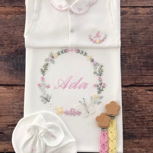 May include: A white baby gown with a pink embroidered collar and a floral wreath design with the name "Ada" in the center. The gown is paired with a white hat and mittens with floral embroidery and two wooden baby teethers with pink and yellow braided cords.