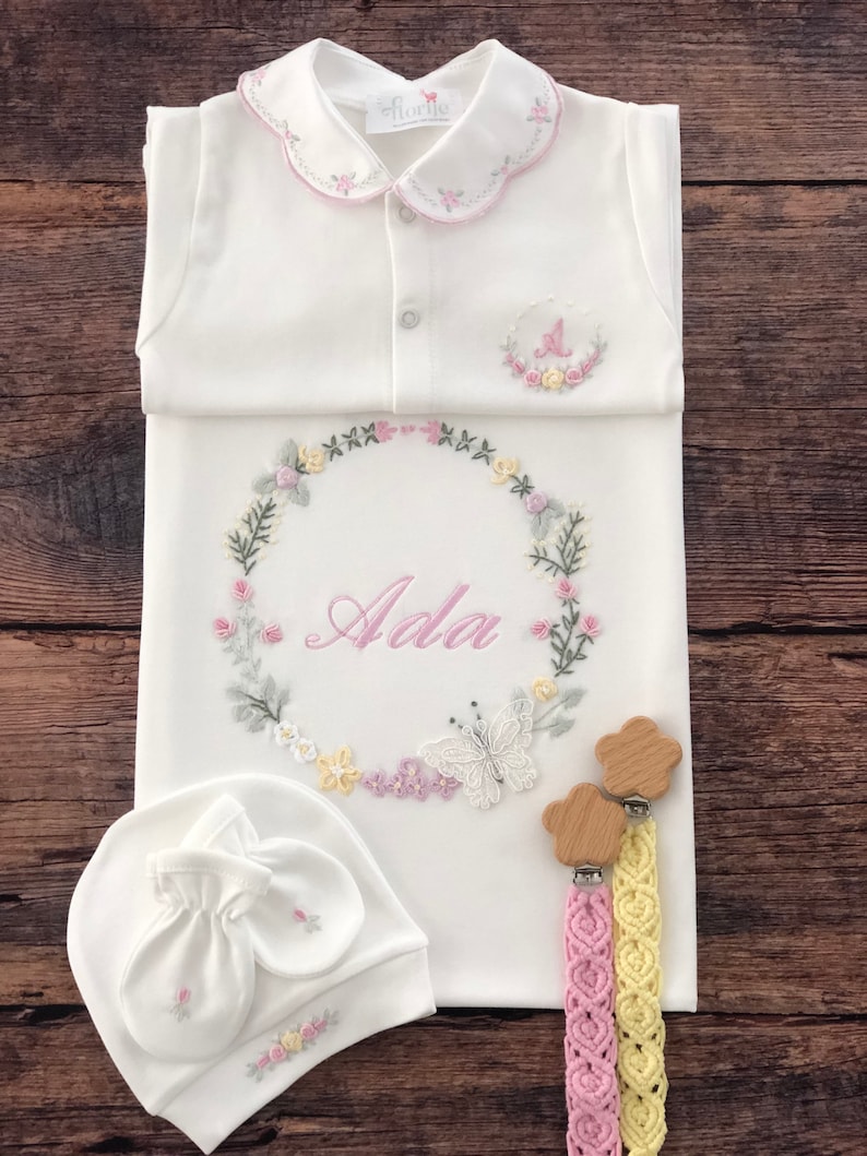 May include: A white baby gown with a pink embroidered collar and a floral wreath design with the name "Ada" in the center. The gown is paired with a white hat and mittens with floral embroidery and two wooden baby teethers with pink and yellow braided cords.