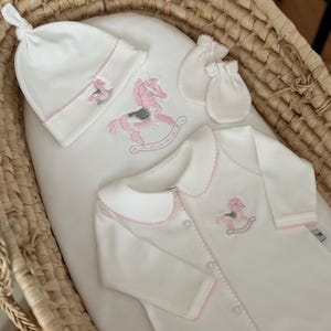 May include: A white baby outfit with a pink rocking horse embroidery. The outfit includes a hat, a onesie, and a pair of mittens.