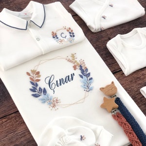 Puede incluir: Un conjunto de regalo para bebé blanco con diseños florales bordados y el nombre "Cinar". El conjunto incluye un body, una camiseta polo, una manta, un par de guantes y un mordedor de madera con una estrella y un gato.