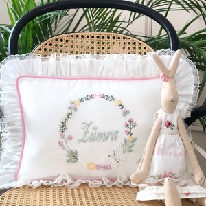 May include: A white pillow with a pink border and a floral wreath embroidered with the name "Zumra" in the center. A white butterfly is embroidered below the wreath. A beige bunny doll with a white dress embroidered with the name "Florije" is sitting on the pillow.