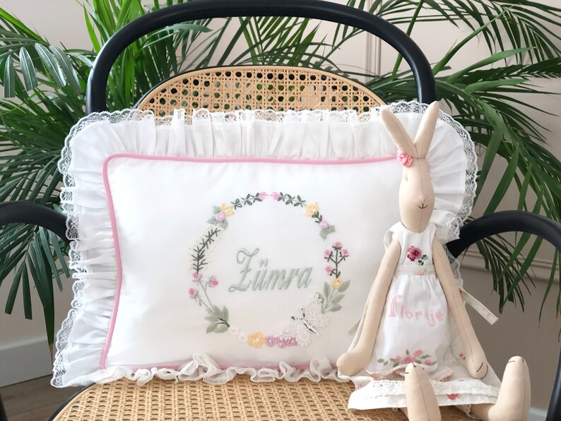 May include: A white pillow with a pink border and a floral wreath embroidered with the name "Zumra" in the center. A white butterfly is embroidered below the wreath. A beige bunny doll with a white dress embroidered with the name "Florije" is sitting on the pillow.