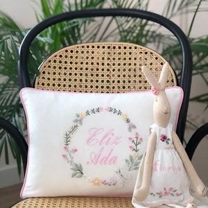 May include: A white pillow with a pink border and embroidered floral wreath with the name "Eliz Ada" in pink lettering. A white bunny doll with a pink dress and the word "Florije" embroidered on it is sitting on the pillow.