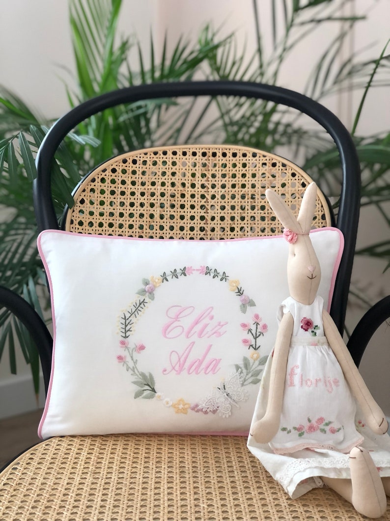 May include: A white pillow with a pink border and embroidered floral wreath with the name "Eliz Ada" in pink lettering. A white bunny doll with a pink dress and the word "Florije" embroidered on it is sitting on the pillow.