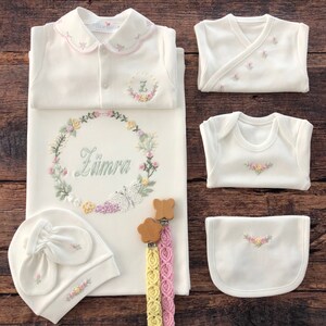 May include: A white baby outfit set with floral embroidery. The set includes a gown, a bib, two bodysuits, a hat, and a pair of wooden teethers with matching ribbon. The gown has a floral wreath with the name "Z&uuml;mra" embroidered on it.