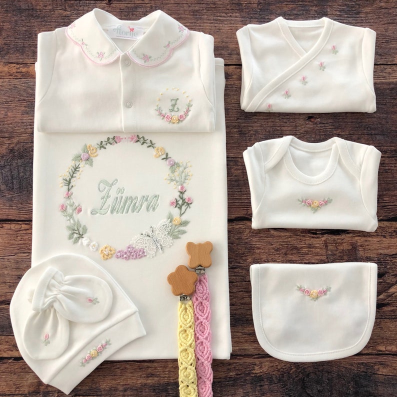 May include: A white baby outfit set with floral embroidery. The set includes a gown, a bib, two bodysuits, a hat, and a pair of wooden teethers with matching ribbon. The gown has a floral wreath with the name "Z&uuml;mra" embroidered on it.