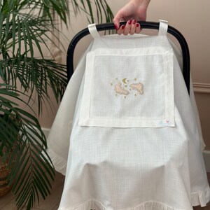 May include: White linen car seat cover with a ruffle trim. The cover features an embroidered design of clouds, a crescent moon, and stars. The text "Little" is embroidered in the bottom right corner.
