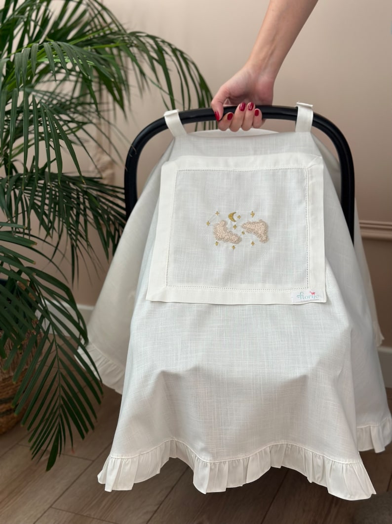 May include: White linen car seat cover with a ruffle trim. The cover features an embroidered design of clouds, a crescent moon, and stars. The text "Little" is embroidered in the bottom right corner.