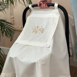May include: White linen baby car seat cover with a ruffle trim. The cover features an embroidered design of two clouds, a crescent moon, and stars. The text "florik" is embroidered in the bottom right corner.