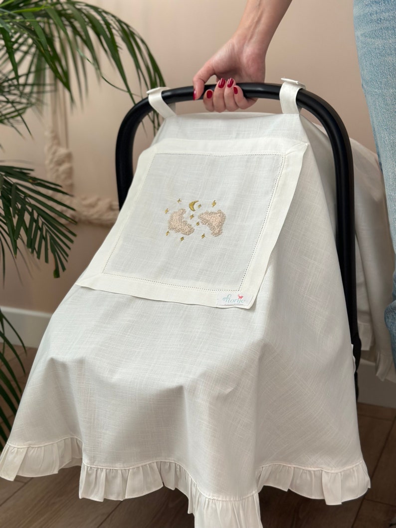May include: White linen baby car seat cover with a ruffle trim. The cover features an embroidered design of two clouds, a crescent moon, and stars. The text "florik" is embroidered in the bottom right corner.