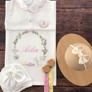 May include: A white baby gown with a pink and green floral embroidery design. The gown has a scalloped collar and a personalized name "Ada" embroidered in the center. The set includes a matching white hat with floral embroidery, a pair of wooden teethers with pink and yellow crocheted straps, and a white headband with a pink bow.