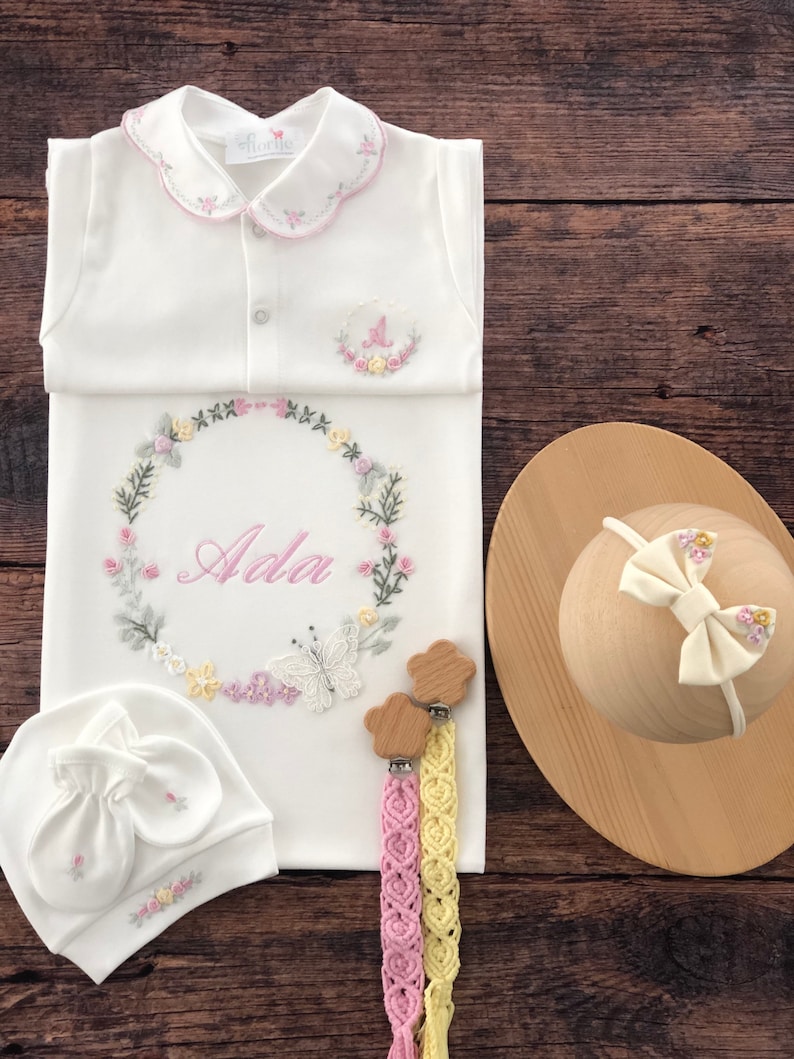 May include: A white baby gown with a pink and green floral embroidery design. The gown has a scalloped collar and a personalized name "Ada" embroidered in the center. The set includes a matching white hat with floral embroidery, a pair of wooden teethers with pink and yellow crocheted straps, and a white headband with a pink bow.