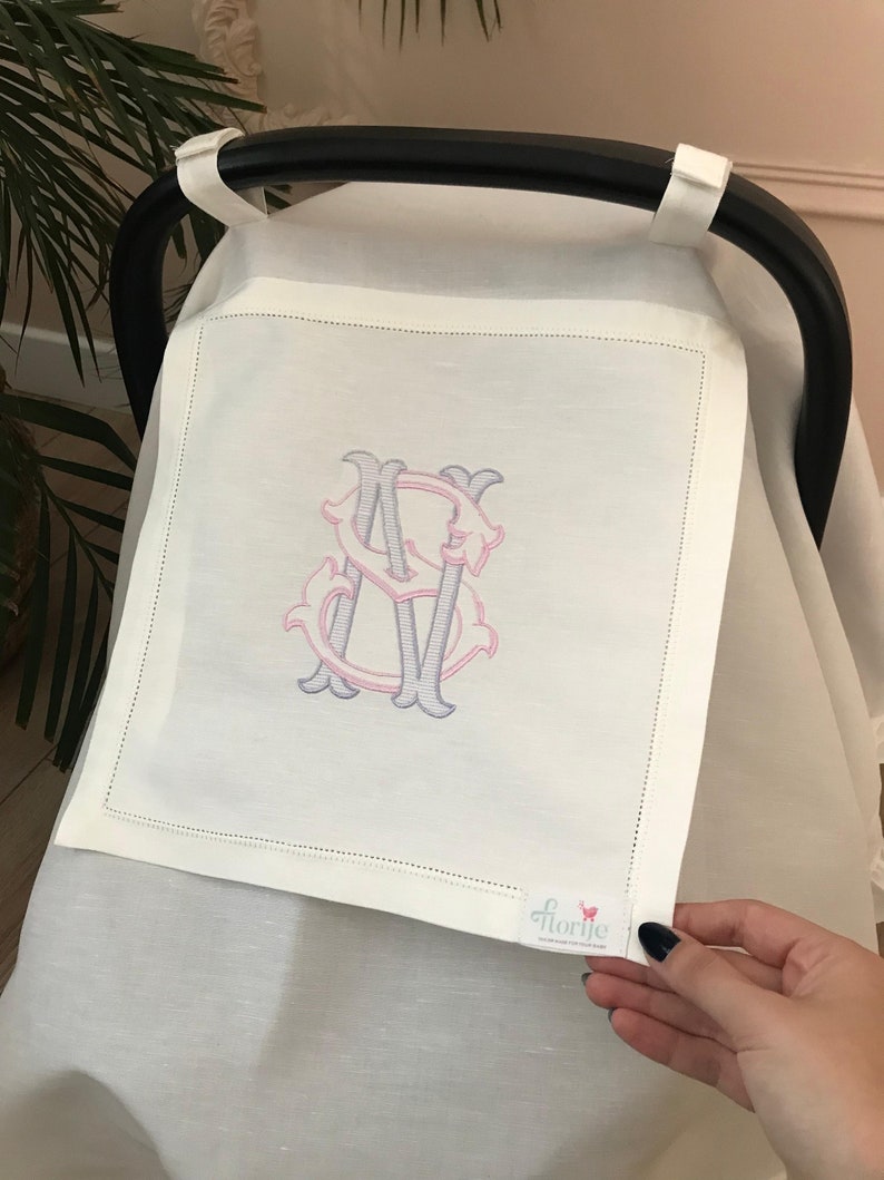 May include: White stroller cover with a pink and purple embroidered monogram "S". The cover has a small white tag with the word "florije" printed on it.