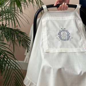May include: White linen stroller cover with a ruffled edge and an embroidered monogram in the center. The monogram is a stylized letter "A" inside a circle.